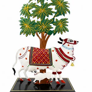 Op de afbeelding: Decoratieve metalen sculptuur met een witte koe met rode en oranje accenten, een kalf en een groene boom met gele accenten. De koe heeft een rood zadel en een bel. De sculptuur staat op een zwarte basis.