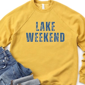 May include: Mustard yellow sweatshirt with the words "LAKE WEEKEND" in blue block letters. The sweatshirt is paired with a pair of blue denim jeans. The clothing is laid out on a white background.