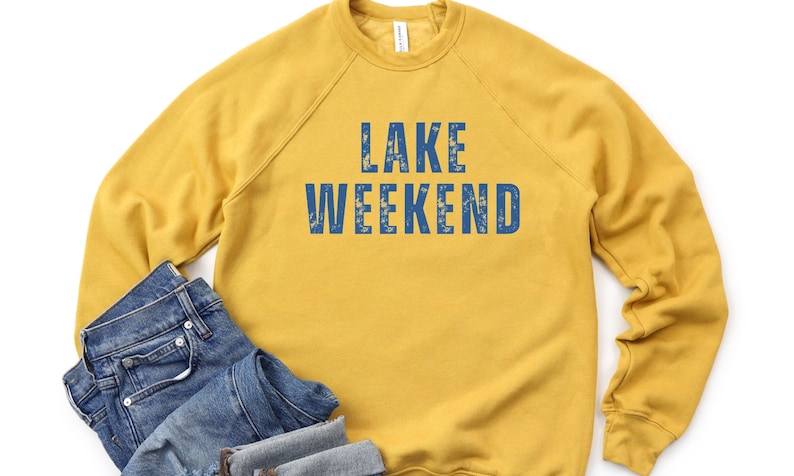 May include: Mustard yellow sweatshirt with the words "LAKE WEEKEND" in blue block letters. The sweatshirt is paired with a pair of blue denim jeans. The clothing is laid out on a white background.