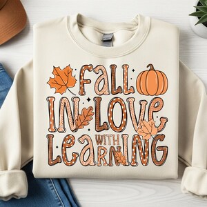 May include: Cream-colored sweatshirt with the text "Fall in Love with Learning" in a fall-themed design. The design includes orange leaves and a pumpkin. A brown baseball cap and blue jeans are also in the image.