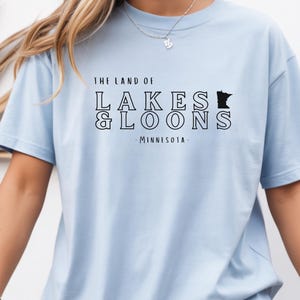 Puede incluir: Camiseta azul claro con el texto negro "THE LAND OF LAKES & LOONS - MINNESOTA". El diseño incluye una silueta del estado de Minnesota. Una prenda de vestir informal.