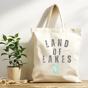 Puede incluir: Bolso tote color crema con el texto "LAND OF LAKES" en gris, con un contorno azul claro de un estado. Una planta en maceta y un rollo de cuerda también son visibles.