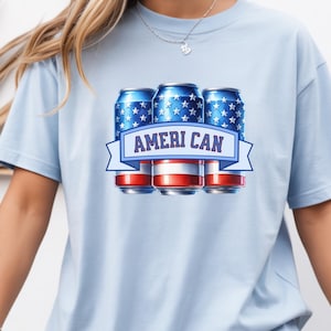 May include: Light blue t-shirt featuring a graphic of three blue cans with white stars, red and white stripes, and the text "AMERI CAN" on a banner. The shirt is a casual style.