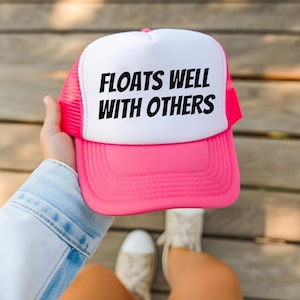 May include: A pink and white trucker hat with the text "FLOATS WELL WITH OTHERS" in black. The hat has a pink brim and mesh back, and a white front panel. The hat is being held up by a person.