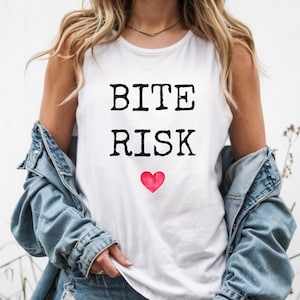 May include: White tank top with the words "BITE RISK" in black, bold letters, and a red heart. The top is worn with a denim jacket.