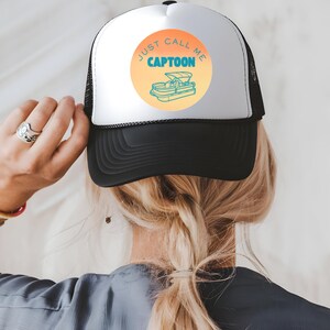 May include: Black and white trucker hat with a white front panel. The hat features a circular design with the text "JUST CALL ME CAPTOON" and a cartoon boat illustration in orange and teal.