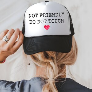 May include: A black and white trucker hat with the text "NOT FRIENDLY DO NOT TOUCH" and a red heart. The hat is worn by a person with blonde hair, and the back of the head and shoulders are visible.