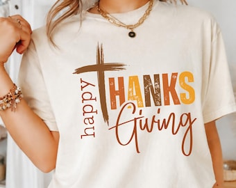 May include: A cream-colored short-sleeve t-shirt featuring a brown cross graphic and the text "happy THANKS Giving" in brown, orange, and yellow. The word "happy" is written vertically, while "THANKS" and "Giving" are in a distressed font.