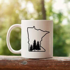 May include: White ceramic mug featuring a black outline of the state of Minnesota with a silhouette of pine trees inside. The mug has a curved handle and is set against a blurred green background.