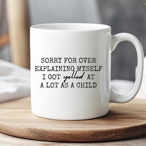 May include: White ceramic mug with the text "SORRY FOR OVER EXPLAINING MYSELF I GOT yelled AT A LOT AS A CHILD" in black. The mug sits on a wooden coaster, with a blurred background.
