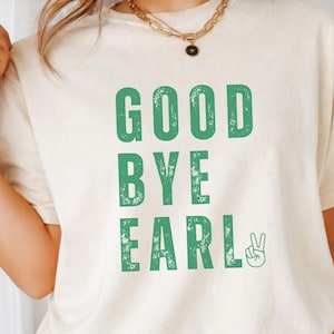 May include: A cream-colored short-sleeve t-shirt featuring the green text GOOD BYE EARL in a distressed, block-letter font. The design includes a small green peace sign hand gesture icon positioned to the right of the word EARL on the chest.