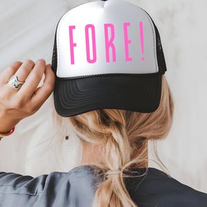 Könnte beinhalten: Schwarz-weiße Trucker-Mütze mit dem Wort "FORE!" in leuchtend pinkfarbenen Buchstaben auf der Vorderseite. Die Mütze hat eine Mesh-Rückseite und einen gebogenen Schirm. Ein lustiges Accessoire für Golf-Enthusiasten.