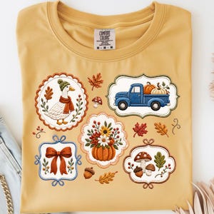 May include: A mustard yellow t-shirt featuring a graphic print of various autumn-themed illustrations including a duck in a green hat and scarf, a blue pickup truck carrying pumpkins, a bow, a pumpkin with flowers, and mushrooms with acorns.