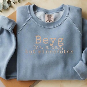 May include: A pale blue sweatshirt featuring the text "Beyg (n.) a bag, but minnesotan" in tan lettering. The sweatshirt is folded, with the sleeves cuffed. The Comfort Colors label is visible.