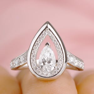 May include: A pear-shaped diamond engagement ring with a halo setting. The ring is set in white gold and features a row of smaller diamonds on the band.