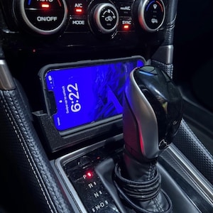 May include: Interior car detail with a smartphone displaying "6:22" on the screen, placed in a console. The image shows the gear shift, climate control buttons, and the "AUTO" and "ON/OFF" buttons. The car's interior is black.