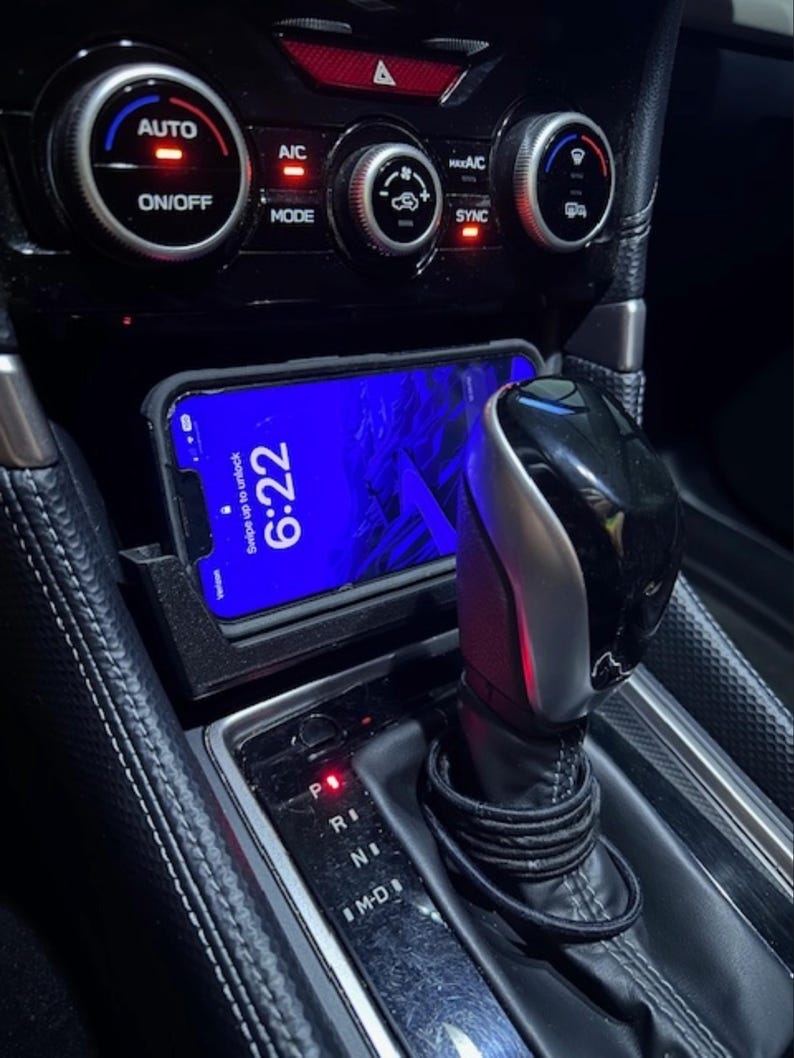May include: Interior car detail with a smartphone displaying "6:22" on the screen, placed in a console. The image shows the gear shift, climate control buttons, and the "AUTO" and "ON/OFF" buttons. The car's interior is black.