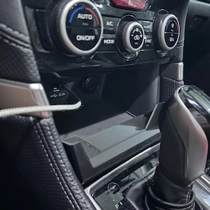 May include: Close-up of a car's interior dashboard featuring climate control knobs, a USB port, and a gear shift. The dashboard is black with silver accents. The climate control knobs have blue and red indicators.