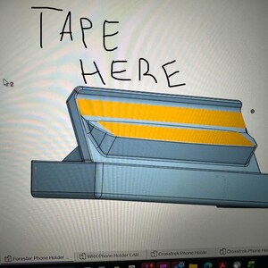 May include: A 3D-rendered phone holder design in shades of gray and yellow. The text "TAPE HERE" is handwritten at the top. The holder has a rectangular base and angled top with two yellow stripes.