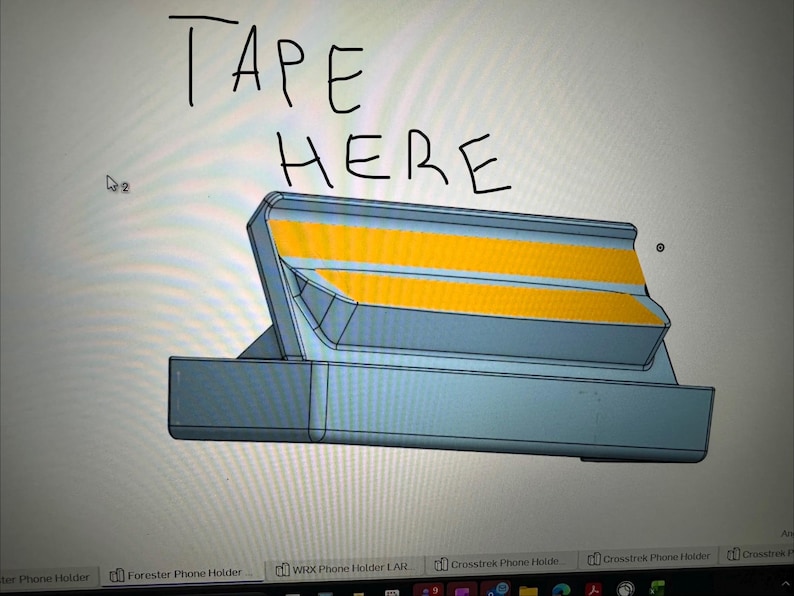 May include: A 3D-rendered phone holder design in shades of gray and yellow. The text "TAPE HERE" is handwritten at the top. The holder has a rectangular base and angled top with two yellow stripes.