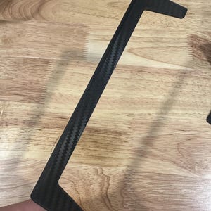 Op de afbeelding: Een zwarte koolstofvezel sierlijst met een kenmerkende L-vorm. De lijst heeft een geweven patroon en wordt tegen een houten oppervlak gehouden. Het stuk lijkt een auto-onderdeel of accessoire te zijn.