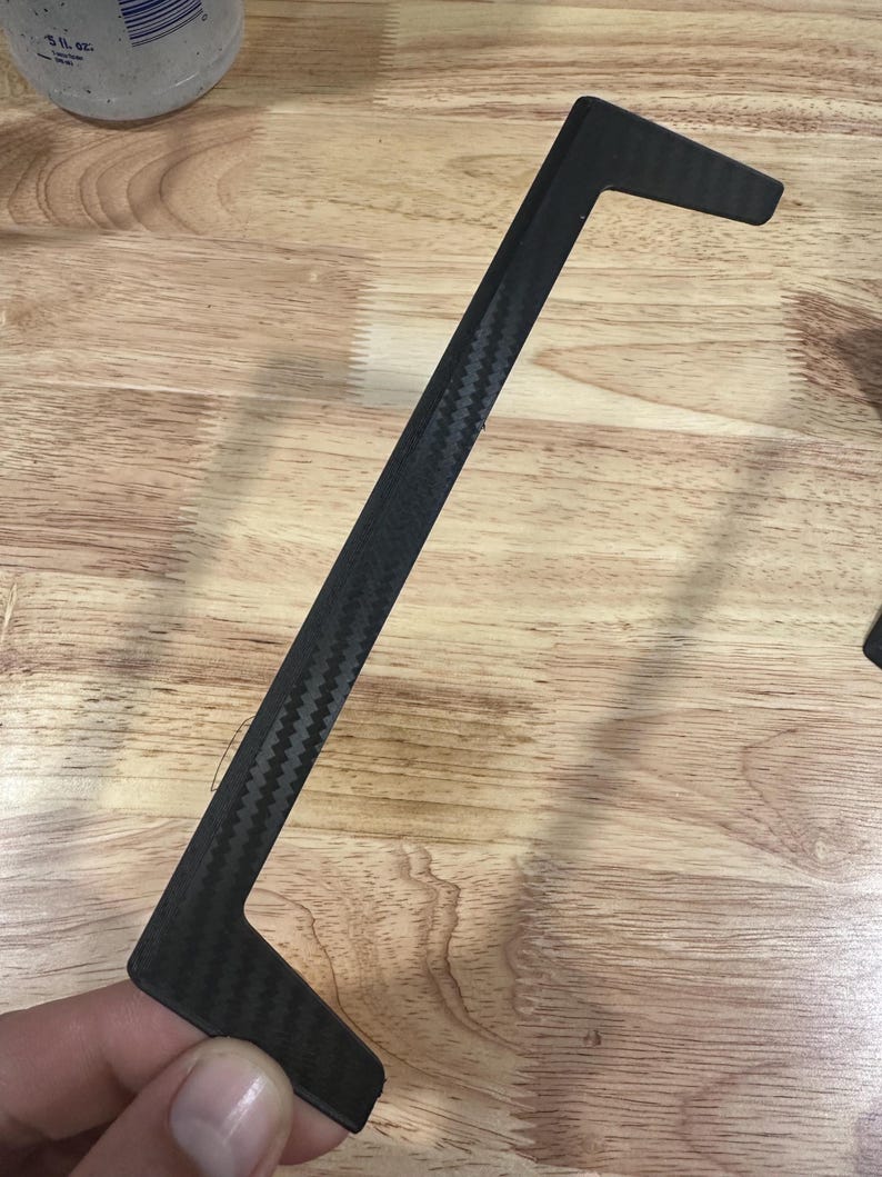 Op de afbeelding: Een zwarte koolstofvezel sierlijst met een kenmerkende L-vorm. De lijst heeft een geweven patroon en wordt tegen een houten oppervlak gehouden. Het stuk lijkt een auto-onderdeel of accessoire te zijn.