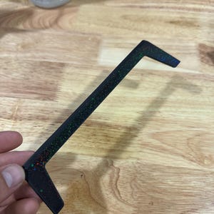 Op de afbeelding: Een zwart, L-vormig object met een gespikkelde, iriserende afwerking. Het object wordt tegen een houten oppervlak gehouden. De gespikkelde afwerking bevat groene, rode en blauwe vlekken. Het object lijkt een decoratief of functioneel stuk te zijn.