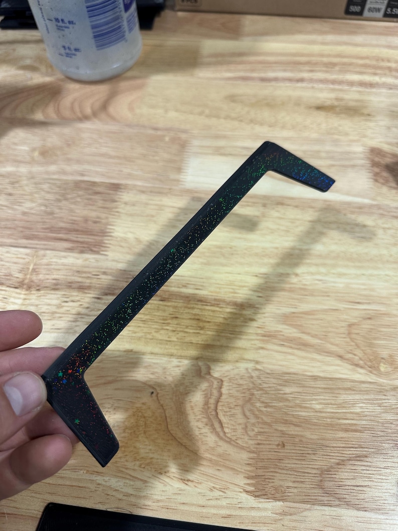 Op de afbeelding: Een zwart, L-vormig object met een gespikkelde, iriserende afwerking. Het object wordt tegen een houten oppervlak gehouden. De gespikkelde afwerking bevat groene, rode en blauwe vlekken. Het object lijkt een decoratief of functioneel stuk te zijn.