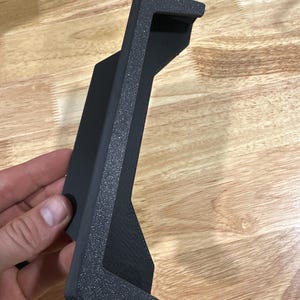 Op de afbeelding: Een zwarte, getextureerde, schuine beugel met een rechthoekige basis en een verhoogde, getextureerde rand. De beugel wordt in een hand gehouden en lijkt gemaakt te zijn van een duurzaam materiaal. Het ontwerp suggereert dat het een steun- of montagecomponent kan zijn.