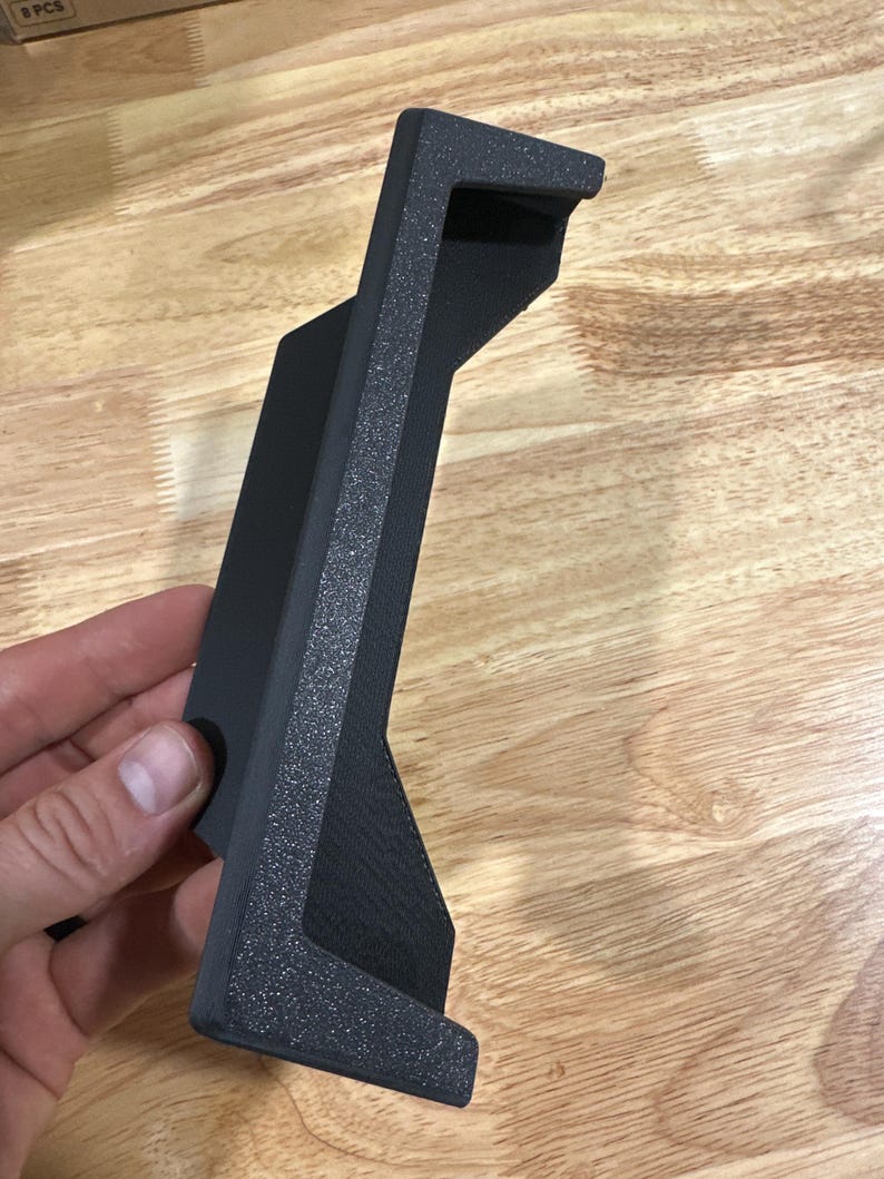Op de afbeelding: Een zwarte, getextureerde, schuine beugel met een rechthoekige basis en een verhoogde, getextureerde rand. De beugel wordt in een hand gehouden en lijkt gemaakt te zijn van een duurzaam materiaal. Het ontwerp suggereert dat het een steun- of montagecomponent kan zijn.