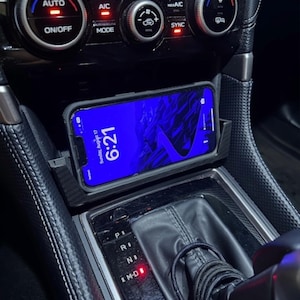 May include: A smartphone displaying "Sunday, August 17, 6:21" rests in a car's console. The phone is in a black case, positioned above the gear shift. Climate control buttons and a gear shift are visible.