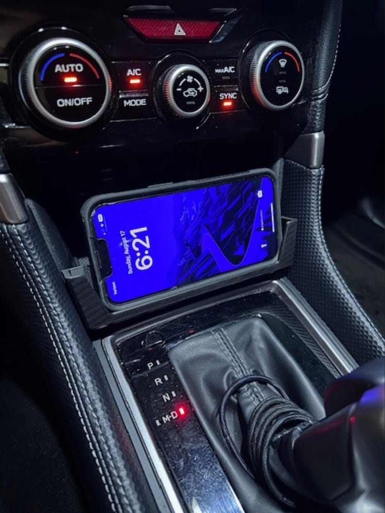 May include: A smartphone displaying "Sunday, August 17, 6:21" rests in a car's console. The phone is in a black case, positioned above the gear shift. Climate control buttons and a gear shift are visible.