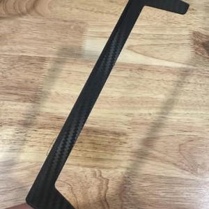 May include: A black, carbon fiber trim piece with a textured weave pattern. The trim has a long, straight section with two right-angled extensions at each end. The object is held against a wooden surface.