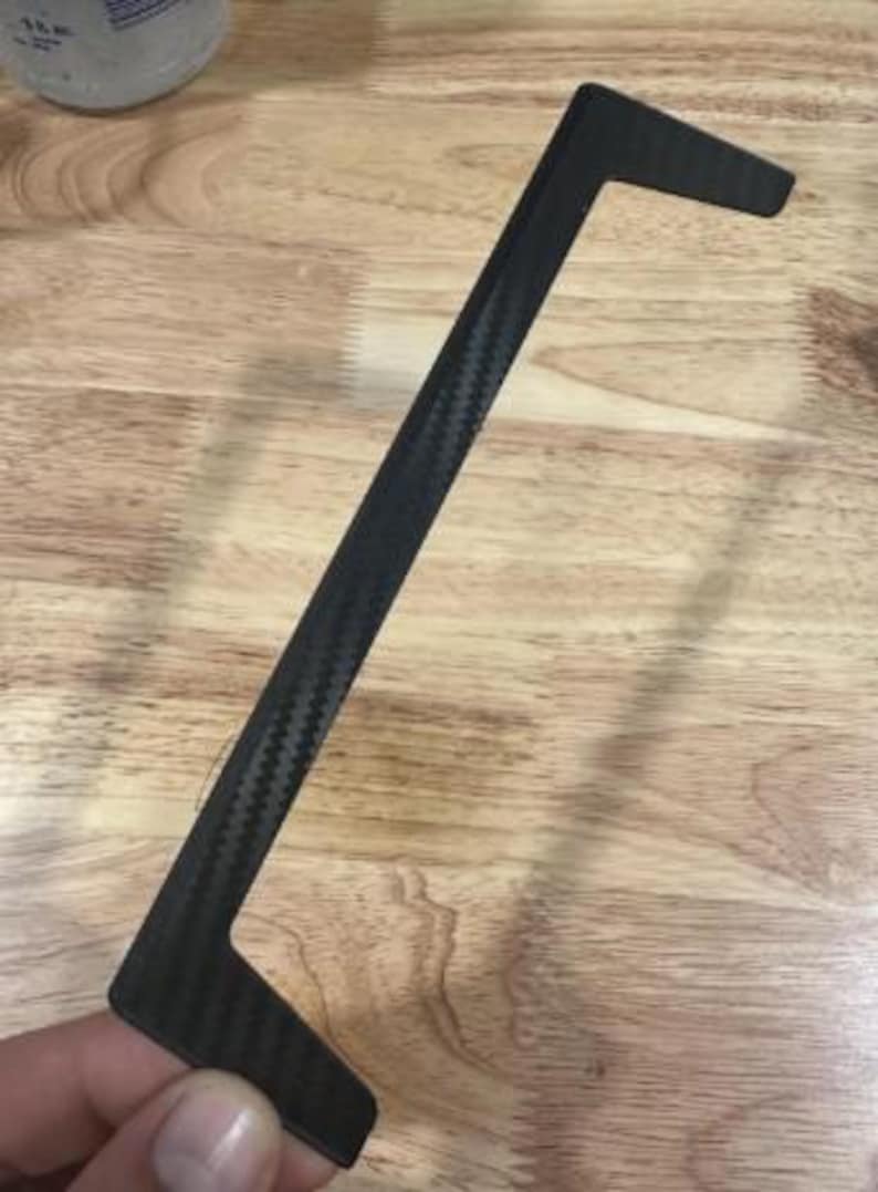 May include: A black, carbon fiber trim piece with a textured weave pattern. The trim has a long, straight section with two right-angled extensions at each end. The object is held against a wooden surface.