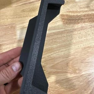 May include: A black, textured, 3D-printed bracket with a rectangular shape and a right-angle bend. The bracket has a textured surface and is designed to hold an item. The bracket is held in a hand.