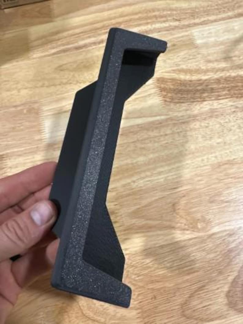 May include: A black, textured, 3D-printed bracket with a rectangular shape and a right-angle bend. The bracket has a textured surface and is designed to hold an item. The bracket is held in a hand.