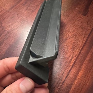 May include: A black, angular plastic object with a textured surface. The object has a right-angle shape with a smaller, attached piece. The item is held in a person's hand.