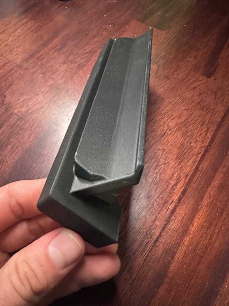 May include: A black, angular plastic object with a textured surface. The object has a right-angle shape with a smaller, attached piece. The item is held in a person's hand.