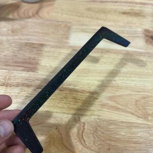 May include: A black, L-shaped object with glitter embedded in the surface. The object is held in a hand and appears to be made of plastic or a similar material. The glitter reflects multiple colors.