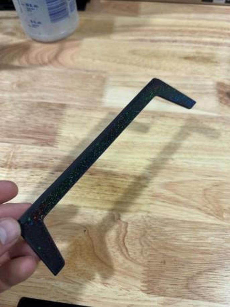 May include: A black, L-shaped object with glitter embedded in the surface. The object is held in a hand and appears to be made of plastic or a similar material. The glitter reflects multiple colors.