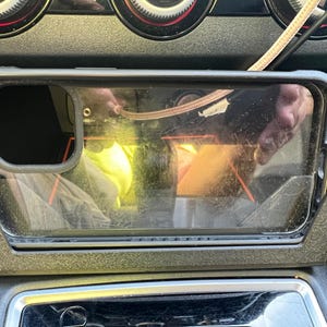 Op de afbeelding: Een doorzichtige telefoonhoes zit in een zwart auto-console. De rechthoekige telefoonhoes heeft een zwarte rand. Een kabel is zichtbaar boven de telefoonhoes.