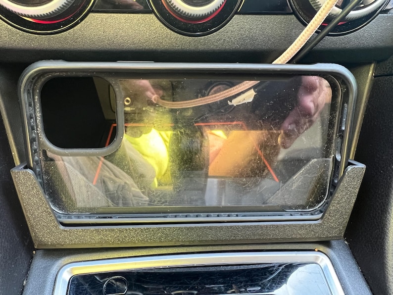 Op de afbeelding: Een doorzichtige telefoonhoes zit in een zwart auto-console. De rechthoekige telefoonhoes heeft een zwarte rand. Een kabel is zichtbaar boven de telefoonhoes.