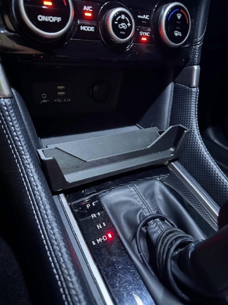 May include: Black car console with a gear shift, a tray, and various buttons and dials. The console includes a USB port and a power outlet. The gear shift is in the 'D' position, indicated by a red light.