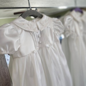 May include: A white satin christening gown with a lace collar and a satin bow at the waist. The gown is hanging on a wooden hanger.