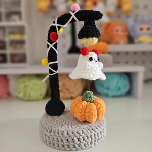 May include: A handmade crochet Halloween decoration featuring a black lamp post with colorful accents and a white ghost. An orange pumpkin sits on a gray crocheted base, creating a festive scene.