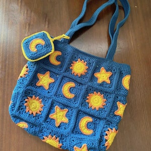 Peut inclure: Un sac fourre-tout en crochet bleu avec une petite pochette assortie. Le sac présente un motif répétitif d'étoiles jaunes, de soleils orange et de lunes jaunes.