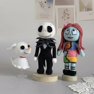 May include: Three handmade crochet dolls featuring a white ghost dog with a red collar, a tall skeleton figure in a black suit with a bat bowtie, and a doll with long red hair wearing a multicolored patchwork dress, all standing on wooden bases.