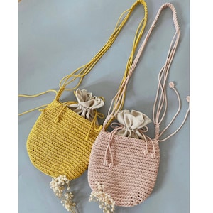 以下が含まれることがあります： 手作りのかぎ針編み巾着バッグ2つ。1つはマスタードイエロー、もう1つはライトピンクです。どちらもベージュの裏地と細長いストラップが付いています。バッグは小さく、必需品を持ち運ぶのに最適です。バッグはライトグレーの表面に表示されています。