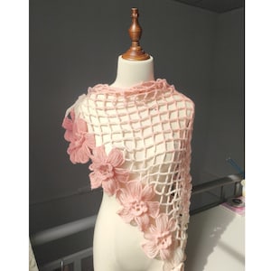 Puede incluir: Un chal de ganchillo hecho a mano, de color crema, con un delicado patrón de red. El chal está adornado con varias flores rosas tridimensionales de hilo a lo largo del borde. El chal está drapeado sobre un maniquí.