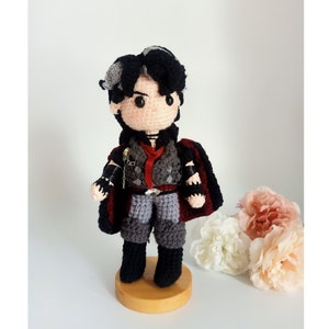 Puede incluir: Juguete de peluche tejido a crochet de un personaje con cabello negro, mechas grises y un atuendo rojo y gris. El juguete está de pie sobre una base de madera.
